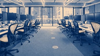 Rows of computer monitors and empty office chairs beside large windows in a blue-toned office.