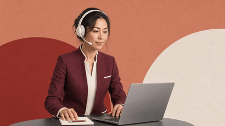 Woman wearing a headset and burgundy blazer working at a laptop against a terracotta and cream background.