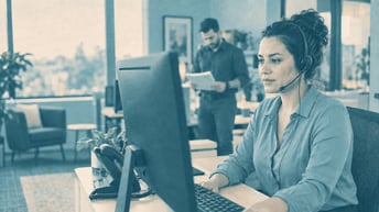 A headset-wearing customer service professional at a workstation while a colleague reviews a report.