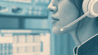 Close-up of a woman in a teal uniform wearing a cream headset, with blurred computer monitors behind her.