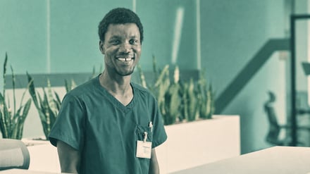 A smiling man in scrubs behind a reception desk, treated in muted teal and warm cream.