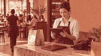 A restaurant manager holding a tablet beside a telephone and takeaway bags during service.