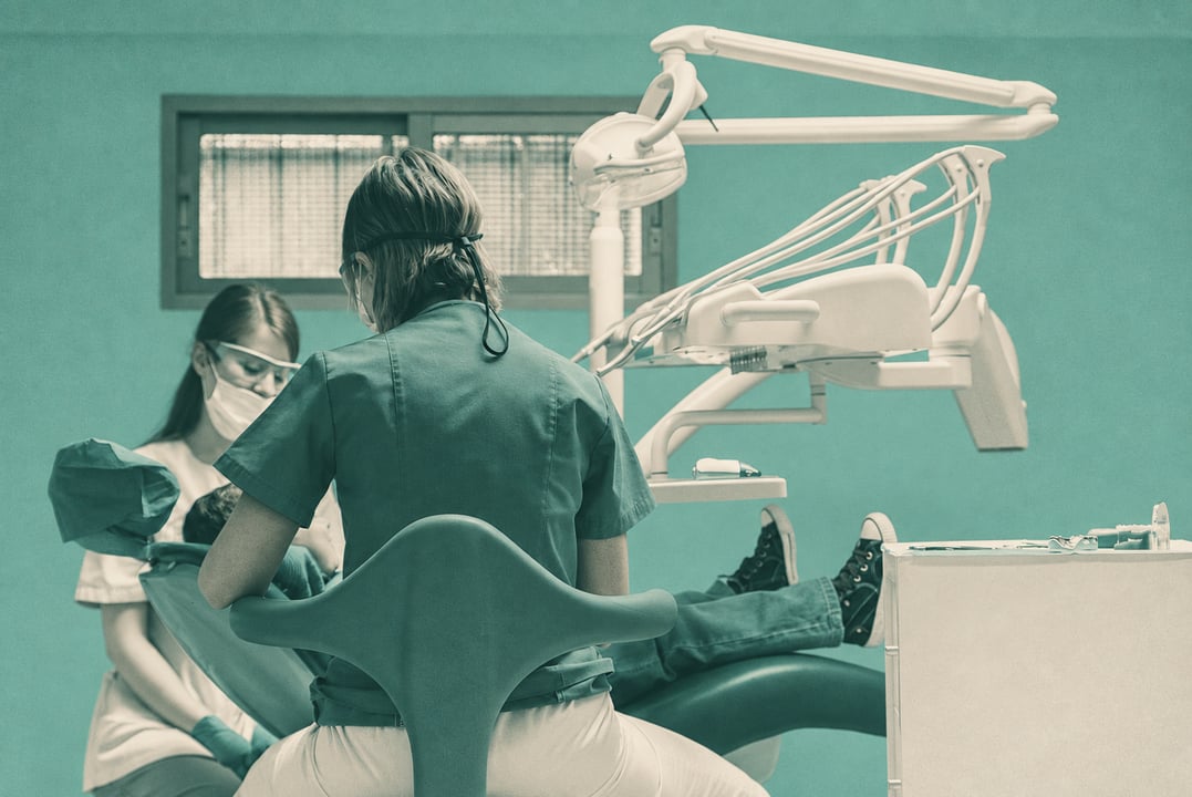 Two dental professionals treating a reclining patient beneath dental equipment in a clinic.